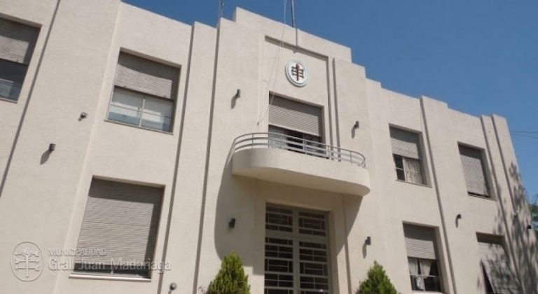 Edificio municipal de Madariaga con balanza desequilibrada, simbolizando la caída de la coparticipación y el riesgo de servicios esenciales.